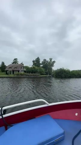 The video shows a first-person perspective from a red and blue boat on a dark, choppy body of water under an overcast sky. The boat is moving forward. On the left bank, there is dense green foliage and a building with a thatched roof. Further ahead, the boat approaches a green, wooden house on stilts with windows, and two ducks are swimming in the water near it. The boat continues to move forward, passing the house.