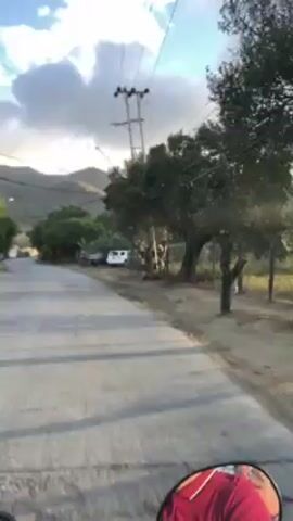 The video depicts a street scene in a semi-rural area with mountains visible in the background. The sky is partly cloudy, and there are power lines running above the street. Several cars are parked on the side of the road. The camera appears to be mounted on a moving vehicle, possibly a motorcycle, as it travels down the paved road. Trees and vegetation line the roadside.
