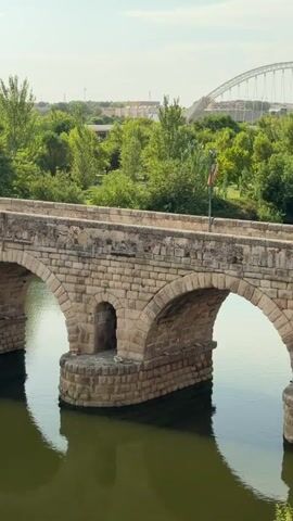 The video shows a stone bridge with multiple arches spanning a body of water. The bridge is constructed from large, rough-hewn stones. There are small, arched alcoves built into the piers of the bridge. Small flags are mounted on poles along the top of the bridge. The water below is a murky green and reflects the arches and piers of the bridge. Trees with lush green foliage line the bank of the water behind the bridge. In the distance, buildings and more trees are visible. The sky is partly cloudy. Birds can be seen flying in the air and some are perched on the bridge. The camera slowly pans across the bridge from right to left.