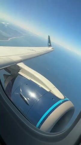 A video captures a vertical perspective from an airplane window, showcasing the wing and engine on a clear, sunny day. The engine is predominantly dark blue with a bright, reflective spot from the sunlight. A light blue stripe runs around its front section. The plane's wing extends from the right, with a small fin at its tip. Beyond the wing, the vast, calm blue ocean stretches, with a distant landmass visible on the left horizon under a clear blue sky. The camera remains static throughout the video, focusing on this high-altitude view.