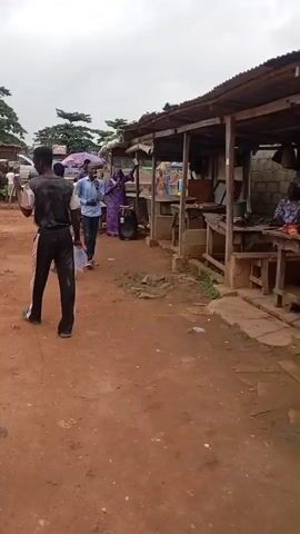 The video shows a dirt road with several people and market stalls. In the foreground, a young man in a black and grey shirt and black pants walks away from the camera, carrying a white plastic bag. To his right, a man in a blue shirt and jeans walks towards the camera, holding a microphone to his mouth and speaking. Behind him, a woman in a purple dress stands near a stall. Further back, a man on a motorcycle is visible, as are several market stalls with colorful umbrellas. The ground is a reddish-brown dirt, and the sky is overcast. The overall impression is of a busy outdoor market.