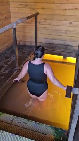 A woman in a black swimsuit is standing in a pool of yellow water. She is holding onto a metal railing and appears to be moving into the pool. The pool is surrounded by wooden walls.
