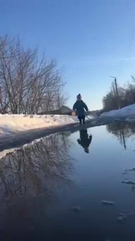A low-angle shot captures a young child running through a puddle on a road. The child, dressed in a green snowsuit and a beanie hat, is running directly towards the camera. The puddle creates a clear reflection of the child and the surrounding environment. Snow is visible on the sides of the road, indicating a winter or early spring setting. Bare trees line the road, and a bright blue sky fills the background. Power lines run across the top of the frame. The overall impression is one of a sunny, possibly cold day with a child enjoying the simple pleasure of splashing in a puddle.