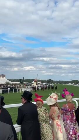 A horse race is underway on a green grassy field under a partly cloudy blue sky. In the foreground, several people are watching the race from behind a white railing. They are dressed in formal attire, including top hats and elaborate hats. The spectators are facing away from the camera and towards the race. In the middle ground, horses and jockeys are running down the track. Various tents and structures are visible in the background, including a white ambulance and a large green crane. The overall scene depicts a formal outdoor event, likely a day at the races.