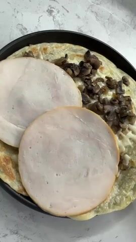 The video shows a close-up, overhead shot of a dark plate with a savory crepe topped with sliced turkey and mushrooms. A red silicone spatula is being used to fold the crepe in half. The crepe appears to be made with chopped green onions. The plate is on a light-colored, textured surface, likely a countertop. The lighting is bright and natural. The video is very short, with the action of folding the crepe taking place over a few seconds.