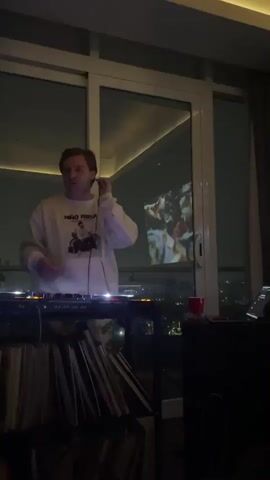 A man with fair skin and light hair, wearing a white long-sleeved shirt with a graphic, stands facing DJ equipment. He is wearing white wired headphones and operating the mixing console, adjusting controls with both hands. Below the DJ equipment, a stack of vinyl records is visible. To the right, a red cup and a black speaker are on a surface. Large windows behind him offer a view of a city skyline at night. A video is projected onto the window glass, displaying what appears to be a crowd of people.