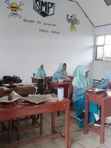 The video depicts a classroom setting with several young female students wearing hijabs seated at desks. They appear to be engaged in an activity, possibly a lesson or a meal, as some have trays or bowls in front of them. The classroom walls are decorated with drawings and text, including a logo that reads "SMP" and a phrase about learning being an investment, not a burden. Sunlight streams in through a large window on the right side of the frame. A teacher, also wearing a hijab, is interacting with students at the back of the classroom. The overall atmosphere appears calm and educational.
