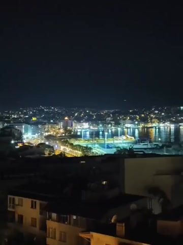 The video shows a nighttime view of a city on a hillside overlooking a body of water with a marina. The city lights are visible, reflecting on the water. The foreground shows the tops of buildings, some with balconies. The sky is dark with a few faint lights, possibly distant aircraft. The scene appears to be from a high vantage point, looking down on the city and harbor. There are no people or animals visible in the video. The video is static, with no camera movement.