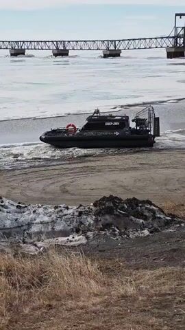 A gray hovercraft maneuvers on a sandy bank near a partially frozen body of water, leaving a wake on the shore. In the background, a steel truss bridge spans the water, and the sky is overcast. The foreground shows a sandy and grassy area with patches of snow. The hovercraft has lettering that reads "CEBEP-2004," followed by an inscription in a different language.