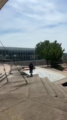 In a daylight scene in Bron, France, a person wearing a black top, patterned trousers, a backpack, and a backward-facing cap is riding a scooter down a sloping, tiled surface. The individual is moving away from the camera, which is positioned higher up, looking down a set of concrete steps.

The tiled area features a mosaic pattern of light blue, white, and grey fragments. In the background, there's a modern building with extensive glass windows, reflecting the bright sky. A large, leafy green tree stands to the right of the building. Construction fencing or similar barriers are visible near the base of the building, suggesting ongoing work or restricted access in that area. The sky is partly cloudy with significant sunlight.