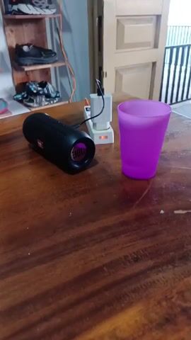 The video opens with a slow pan across a wooden table in an indoor setting. A black, cylindrical JBL portable speaker is positioned horizontally on the table. One end of the speaker emits a distinct purple light. It is connected by a black cable to a white USB power adapter, which is plugged into a white power strip.

Next to the speaker, on the right, rests a translucent purple plastic cup. As the camera continues to pan left, an olive green coiled cable and a small green-handled screwdriver are visible on the wooden surface.

In the background, a wooden shelving unit displays several pairs of athletic shoes in various colors and styles. Further back, a light brown wooden door is partially visible, beyond which a white slatted fence or grill can be seen. The scene depicts a casual, everyday environment, possibly a room in a home.