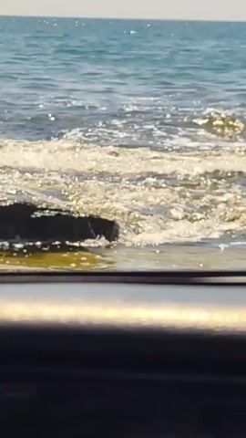 The video shows a close-up view of the ocean from a car, looking out through the windshield. The water is a clear blue, with small waves breaking against dark rocks on the shore. Sunlight glints off the water, creating a shimmering effect. The foreground of the video is blurred, but it appears to be the interior of a car, with the side mirror visible on the left side of the frame. The sky is clear and blue, with no clouds visible. The overall scene is peaceful and serene, evoking a sense of calm and tranquility.