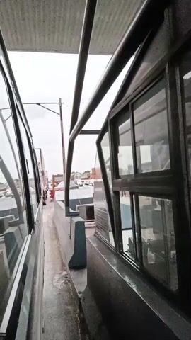 The video shows a view from a vehicle at a toll plaza. A toll booth is visible on the right, with a barrier arm that is partially raised. A man in a yellow safety vest is standing near the toll booth. Several trucks are lined up in the lanes, and traffic appears to be moving slowly. The toll plaza structure is overhead, with signs in English and another language that is not clearly identifiable. The environment appears to be outdoors, with an overcast sky.