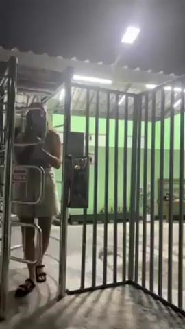 A person walks through a gated turnstile. The person, who appears to be female, is wearing a light blue top, khaki shorts, and dark sandals. She also has a bag slung across her torso.  She pushes through the turnstile to the left of the gate.  The gate is made of metal bars and has a payment mechanism attached to it. Behind the gate is a green wall and some plants. The lighting is dim, but there is an overhead light illuminating the area.