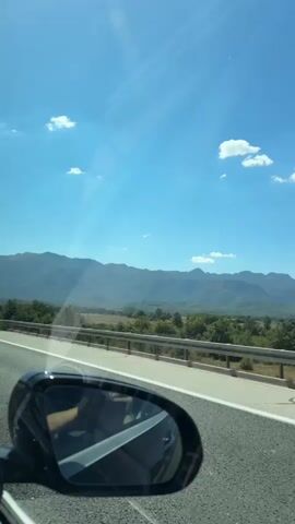 The video shows a view from inside a car, looking forward and slightly to the side, through the side mirror. The car is on a highway, with a road and a guardrail visible in the foreground. In the distance, there are mountains, trees, and a clear blue sky with a few white clouds. Sunlight is shining brightly, casting reflections on the car's window and the side mirror. A road sign indicating directions to Gračac, Lovinac, and Sveti Rok is visible overhead. The car appears to be moving.