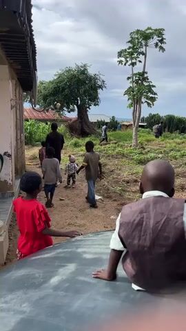 The video shows a group of people, including adults and children, walking along a dirt path. In the foreground, two children are seated on what appears to be the hood of a car, with their backs to the camera. One child is wearing a red top, and the other is wearing a brown vest over a white shirt. The path leads towards a rural landscape with houses, trees, and vegetation. Several other people are visible walking away from the camera further down the path and in the background. The sky is overcast. There is a large tree with a twisted trunk on the left side of the path. Objects are visible in the air near the tree, possibly being thrown. The ground is dirt with some scattered debris.