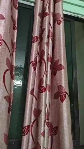 A curtain with a floral pattern is shown. The curtain is light pink with dark red flowers and vines. The curtain is partially covering a window, and a sliding door can be seen behind it. The room appears to be dimly lit.