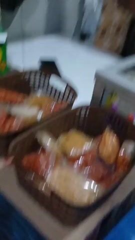 The video shows two plastic containers filled with food. A person is holding one of the containers, which appears to contain some kind of frozen food or drink that is beginning to melt. The person is speaking in Indonesian and the other person is responding. The background is blurry but appears to be an indoor environment.