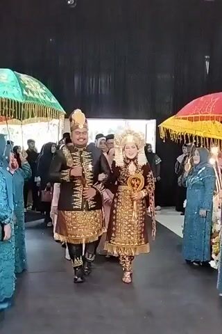 A couple, dressed in traditional ceremonial attire, walks forward, arm in arm. The man wears a dark, elaborately decorated jacket and skirt-like garment with gold accents and a tall headdress. The woman is adorned in a gold and deep red outfit, also richly decorated, with a striking, tall gold headdress. They are walking down a pathway between two rows of people. Several individuals on either side hold colorful, ornate umbrellas above the couple. The background appears to be an indoor event space with dark drapery. The overall scene depicts a formal procession, likely a wedding.