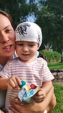 A woman and a young child are in an outdoor setting. The woman is on the left, smiling and looking at the camera. She has short brown hair and is wearing a gray tank top. The child is on the right, also looking away from the camera and smiling. The child has brown hair and is wearing a white bandana with a black design and a striped t-shirt. The child is holding a small green object and a pouch with images of fruit and berries. They are surrounded by greenery, with trees and grass visible in the background. The lighting suggests it is daytime.