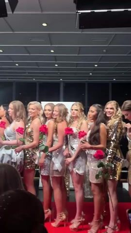 A beauty pageant is in progress indoors. Approximately 20 young women stand shoulder to shoulder on a red-carpeted stage, facing the camera. Each woman holds a red rose. They are wearing short sequined or glittery dresses in shades of gold, silver, and white, and most are wearing heels. Several of them have sashes with writing on them. As the camera zooms out, the setting is revealed as a stage with a backdrop of white draped fabric, floral arrangements, and uplighting. A person in the middle of the group wears a red gown, a crown, and a sash that reads “Miss Denmark 2023”. People in the audience are holding up phones to record the event. The lighting in the hall is bright from stage lights. Overhead, the ceiling structure is visible with track lighting.