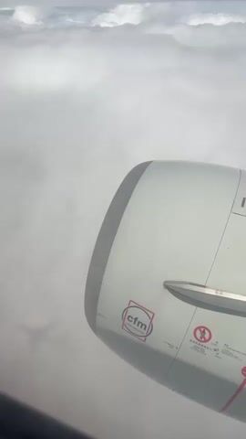 The video shows a view from an airplane window looking down at clouds. Part of an airplane engine is visible in the foreground on the right side of the frame. The CFM logo is visible on the engine. The plane is flying above a thick layer of white clouds, with patches of blue sky visible higher up. The shadow of another airplane wing is visible in the clouds on the left side of the frame. The video appears to be taken while the plane is in flight.