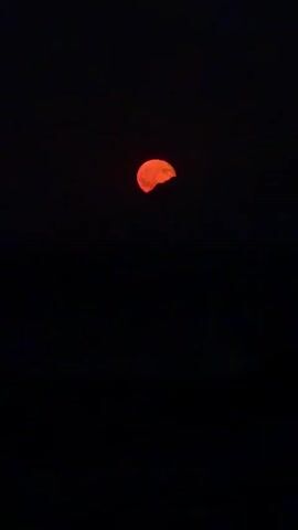 A video shows a partial view of a blood-orange moon against a black sky. The moon is partially obscured by what appears to be darkness or clouds, giving it a jagged appearance. The bottom half of the video is completely black, suggesting a dark environment or possibly the sea. There are no people or other discernible objects visible.