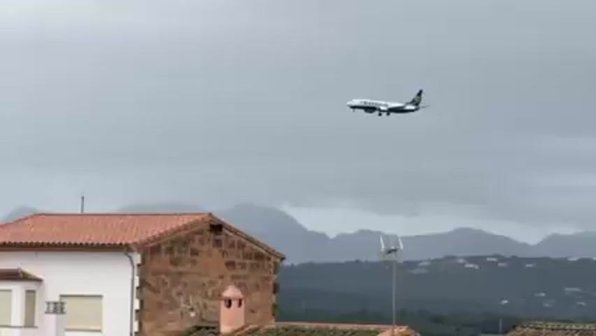 A Ryanair plane is seen in the sky, approaching for landing. The plane is flying in front of a view of houses and distant mountains. The sky is cloudy. The plane has its landing gear down.