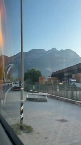 The video shows a view from a vehicle moving along a road. The scene is dominated by a mountain range in the background under a clear blue sky. In the foreground, there are several vehicles and objects visible. On the right side of the road, a flatbed truck carrying numerous wooden pallets is parked. Further down the road, another truck with a trailer is seen. A tow truck with an orange and white body is parked closer to the viewer on the left side of the road. Reflections on the vehicle's window are noticeable, including what appears to be part of the vehicle's interior and possibly a side mirror. The area appears to be an industrial or storage yard adjacent to the road, with a fence separating it from the road.
