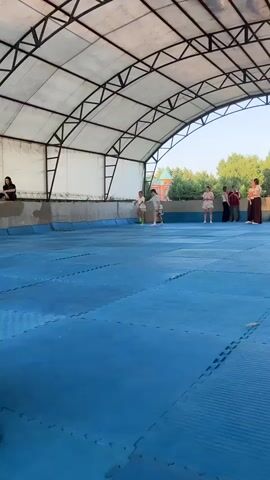 Two young people, one in a white t-shirt and shorts with green shoes, and another in a grey t-shirt and shorts, appear to be practicing some kind of martial art on a blue mat under a covered outdoor area. They are facing each other, and the person in grey is in a low stance, possibly a defensive or attacking posture. In the background, four other young people are standing, watching. One person is wearing a pink t-shirt and shorts, another a black t-shirt and pants, and two are wearing what appear to be red shirts. A building with a distinctive green roof is visible behind them, surrounded by trees. The ground is covered with blue interlocking mats.