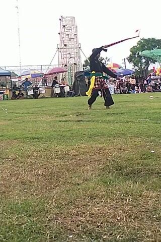 Here is a description of the video:

The video shows a person, possibly male, dressed in a black top, black pants, and a head covering performing with a long, flexible whip or rope-like weapon. The performer is on a grassy field, with a crowd of people in the background, partially obscured by tents or umbrellas. Behind them, there is what appears to be a tall, open framework, possibly part of a carnival ride or stage structure. The performer swings and manipulates the whip in what appears to be a display of skill or a performance routine. The video is rotated on its side.