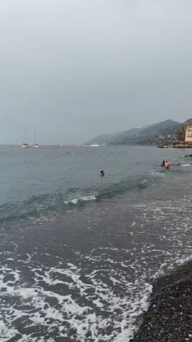 The video shows a choppy ocean with a dark, pebbly beach in the foreground. There are several boats in the distance, including sailboats and a larger vessel. Two people are swimming in the water, one of whom appears to be a child with an adult. The sky is overcast, suggesting potentially inclement weather. The waves are crashing onto the shore, creating white foam. The coastline curves into the distance, with hills or mountains visible behind it.