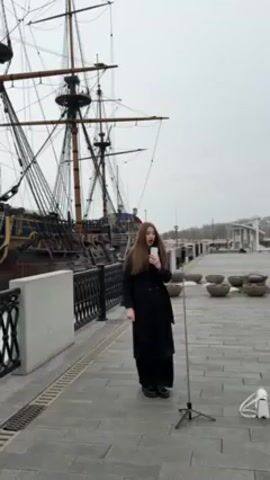 A woman stands on a paved walkway, speaking into a smartphone held in her hand. The phone appears to be on a selfie stick tripod next to her. She is wearing a long black coat and dark pants and shoes. Behind her is a large, dark-colored sailing ship with multiple masts and rigging. The sky is overcast, and the walkway is bordered by a decorative fence and a row of concrete planters. A white purse is visible on the ground to the right of the woman.