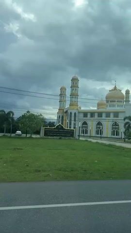 The video shows a large, ornate mosque with multiple domes and minarets. The mosque is predominantly white with gold accents. It is situated in a grassy area with some palm trees. A paved road runs in front of the mosque, and a darker asphalt road is visible in the foreground, suggesting the camera is in a vehicle moving past. The sky is cloudy and overcast. There are a few people walking in front of the mosque and a white car parked on the grounds. A sign is visible to the left of the mosque. The camera appears to be positioned at a slight downward angle.