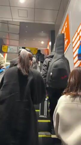 The camera is positioned on an ascending escalator, capturing the backs of several individuals. One person has a black guitar case on their back. Other people are wearing a variety of winter-type clothing. The escalator is in a building with white and grey walls. A yellow sign with arrows is visible on the wall to the left, and an orange advertisement is visible on the wall to the right.