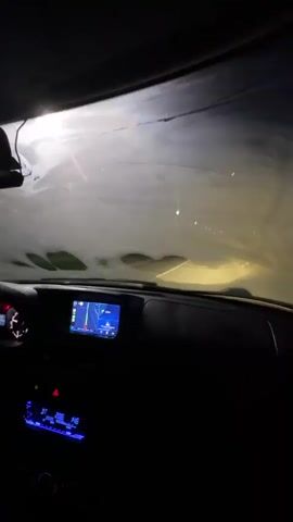 The video shows the interior of a car driving at night. The windshield is covered in snow and ice, obscuring the view of the road ahead. A navigation system is visible on the dashboard, displaying a map with a route. The car's interior lights are on, casting a blue glow on the dashboard. The driver's side air vent and part of the steering wheel are visible. The scene is dark, with the headlights of the car illuminating the snowy road.