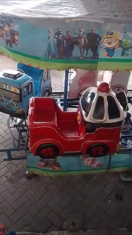 The video shows a children’s ride with four small, brightly-colored vehicles attached to a rotating platform. The vehicles are shaped like a blue bus, a pink carriage, a red car, and a green car. The platform is decorated with cartoon characters. A canopy covers the ride, featuring images of various animated characters, including characters from “Frozen” and “Tayo the Little Bus”. The ride is set up on a paved surface.