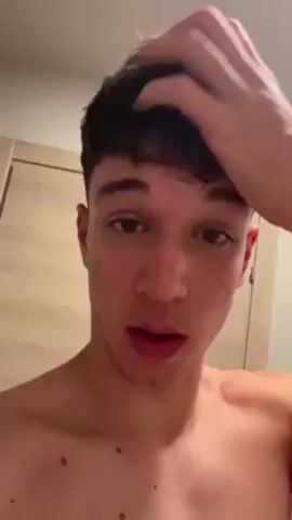 A young man with curly brown hair stands shirtless in what appears to be a bathroom. He looks directly at the camera and runs his hand through his hair. The background is a plain light-colored wall with a door.