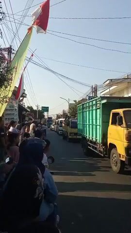 A traffic jam is visible on a sunny day. Several trucks and cars are parked on the street, with people standing on the side of the road. The Indonesian flag is flying on a pole, and there are decorations hanging overhead. The scene appears to be a town or city street.