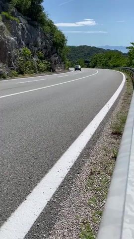 A black Mercedes-Benz C-Class Coupe drives from right to left on a curved asphalt road. The road has a white solid line on the right shoulder and a white double solid line in the middle. Green trees and a rock formation are visible on the left side of the road, and green trees and mountains are visible on the right. A metal guardrail is on the right side of the road. The sky is blue and clear.