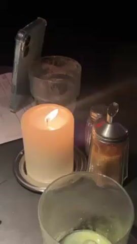 The video shows a dimly lit table setting with a lit candle as the primary light source. A smartphone in a clear case leans against a dirty glass. Beside the glass are a salt shaker and a sugar dispenser.  Another empty glass is in the foreground. The surface appears to be a dark, possibly stone, table. A white bill or receipt is partially visible behind the phone.