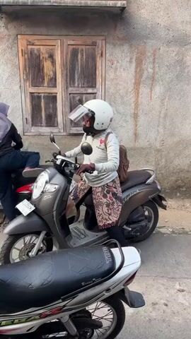 This video shows a street scene with several motorcycles parked. A person wearing a white helmet and a light-colored top with a patterned skirt is sitting on a motorcycle. Another person in a hijab and dark clothing is standing next to a motorcycle. A yellow motorcycle is parked nearby, and a person in a red top and striped pants is standing next to it. Further down the street, there is a building with a wooden-framed window and a wooden door. A red motorcycle is parked in front of the window. Another motorcycle, black and red, is parked next to it. A person wearing a pink hijab and a patterned dress, with a young boy wearing a red and white shirt and a backpack, are sitting on a white motorcycle. More motorcycles are parked along the street. The environment appears to be an outdoor, possibly urban or suburban, setting.