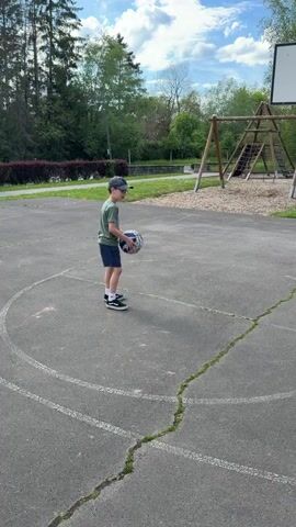 The video shows a child shooting a basketball. He is standing on an asphalt basketball court. There is a basketball hoop and backboard in front of him. There is a children's swing set and climbing structure in the background. The child bounces the ball a few times, throws it at the hoop, and misses. The sky above is blue with scattered white clouds.