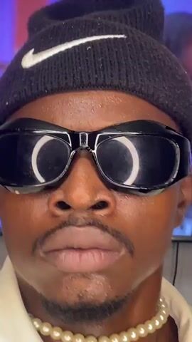 A close-up shot of a person's face wearing a black beanie with a white Nike logo, black wraparound sunglasses, and a pearl necklace. The person has a mustache and is looking directly at the camera. The background is blurred, but appears to be an indoor setting with some colorful lights. The person's facial expression is neutral. There are no discernable actions or interactions. The video is very short, lasting only a few seconds.