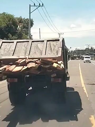 A dump truck loaded with irregularly shaped planks of wood is driving on a road with a yellow lane divider. The truck is gray and its rear gate has four large horizontal rectangular panels. The wood appears to be overflowing from the bed of the truck. Power lines run overhead. The weather is sunny. Other cars can be seen in the distance. The camera appears to be in a following vehicle. A loud crashing or splintering sound is audible.