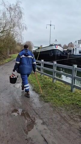A person in a blue uniform, holding a black bucket, walks away from the camera on a muddy path alongside a canal. The canal has a grey and white wooden fence bordering it. A large boat, named "SHANGHAIOL", is docked in the canal. Bare trees line the opposite side of the path. The sky is overcast. The person also has a yellow object in their other hand.