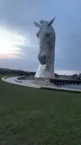 The video shows two large, silver horse head sculptures known as the Kelpies. They are situated in a body of water, with a grassy area in the foreground. The sky is overcast, with a hint of light on the horizon, indicating dusk or dawn.