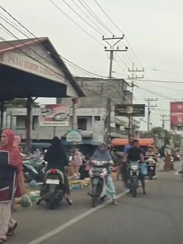 The video shows a busy street scene in what appears to be a market area. There are many people, most of whom are on motorcycles. The people are dressed in casual clothing, and many of the women are wearing headscarves. The motorcycles are parked along the side of the road, and some people are walking between them. There are also market stalls set up along the side of the road, with vendors selling goods. The overall atmosphere is one of activity and commerce.