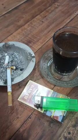 The video opens with a close-up of an ashtray containing cigarette butts and ashes, a pack of cigarettes, a green lighter, and a glass of coffee on a wooden table. The perspective then shifts, showing a person from behind, wearing a light-colored long-sleeved shirt and a dark baseball cap, sitting at the same wooden table. The camera pans to the right, revealing more of the table and a second glass of coffee. The background shows an outdoor setting with motorcycles parked nearby, a palm tree, and buildings. The lighting suggests it is late afternoon or early evening. The person remains seated throughout the recorded sequence, facing away from the camera.