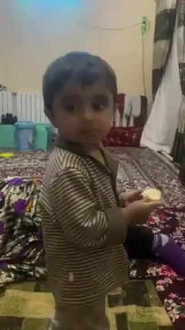 A young child with dark hair is standing and holding a light-colored object in their hands, possibly food. The child is wearing a striped brown and tan shirt and patterned pants. They are standing on a patterned rug in a room with a bed or large cushion covered in a similar patterned fabric. In the background, there is a white radiator, a green plastic container, a curtain with brown and silver vertical stripes, and some hanging clothes. The lighting appears to be indoor and possibly artificial.